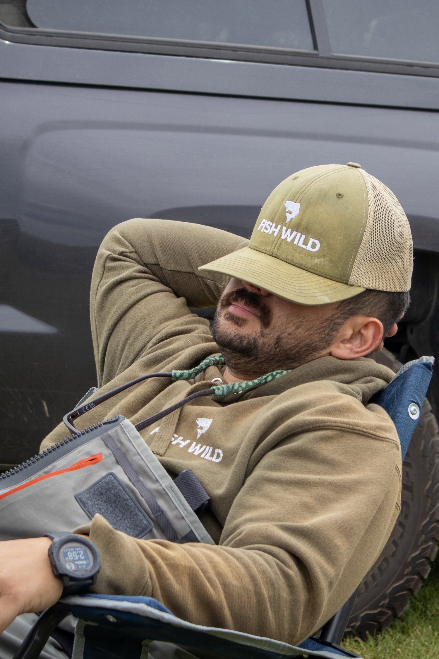 Man sitting in a chair next to a truck wearing a cap with a Fish Wild logo